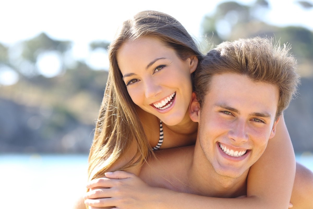 Young couple smiling outdoors by water in sunny setting, promoting dental care wellness.