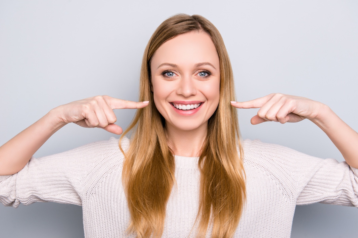 Smiling woman pointing to her teeth against gray background, promoting dental care.