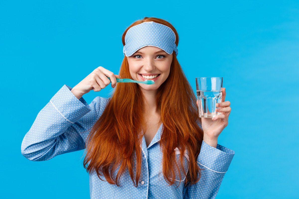 Woman brushing teeth in pajamas holding glass of water against blue background.