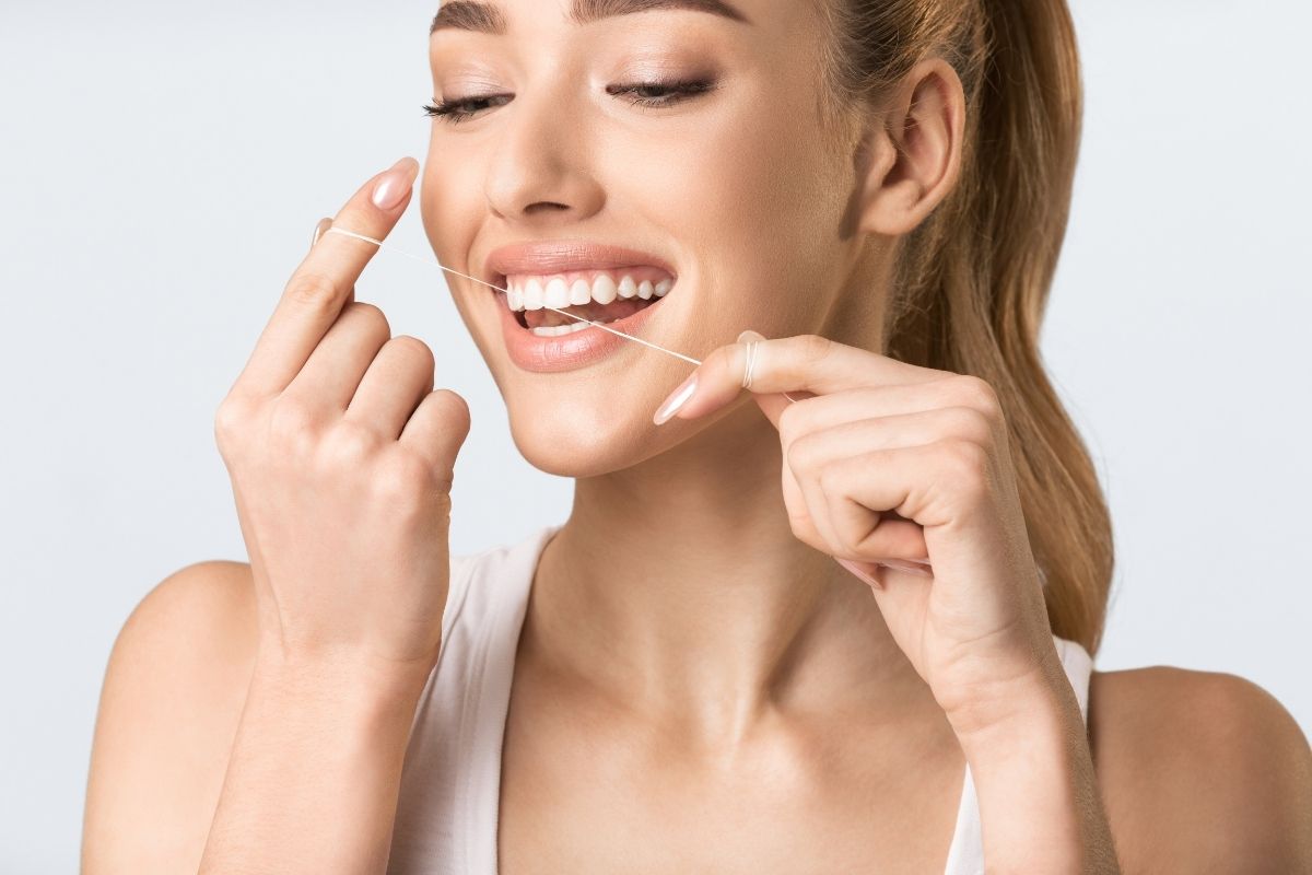 Woman flossing teeth with white dental floss, smiling against neutral background.
