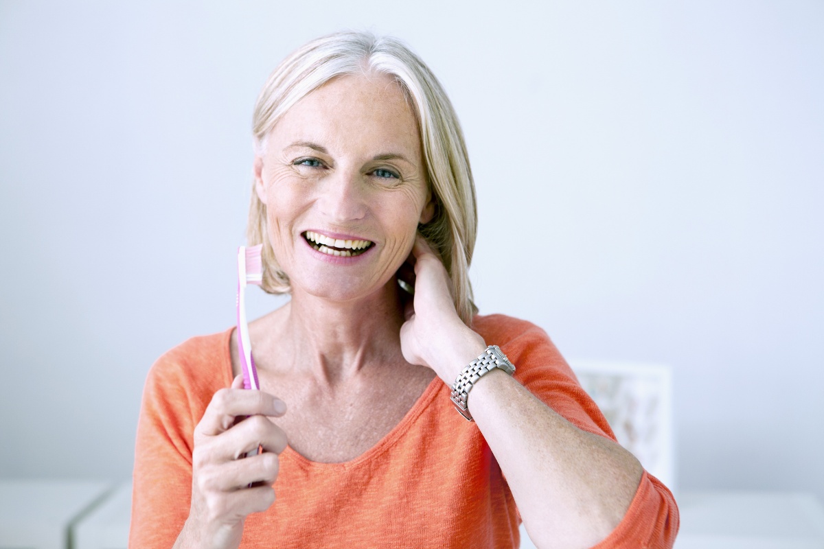 Smiling mature woman in orange shirt touching face, bright indoor setting for dental care website.