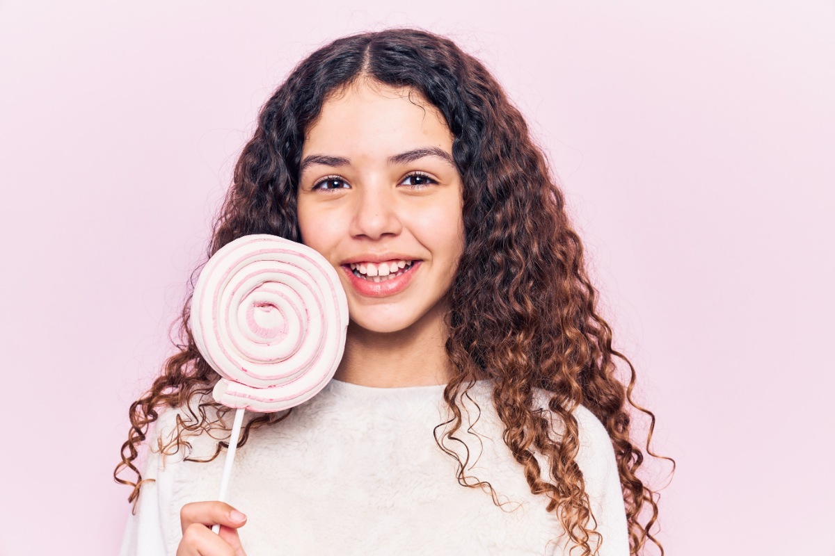 Smiling woman with curly hair holding large decorative tooth model against pink background