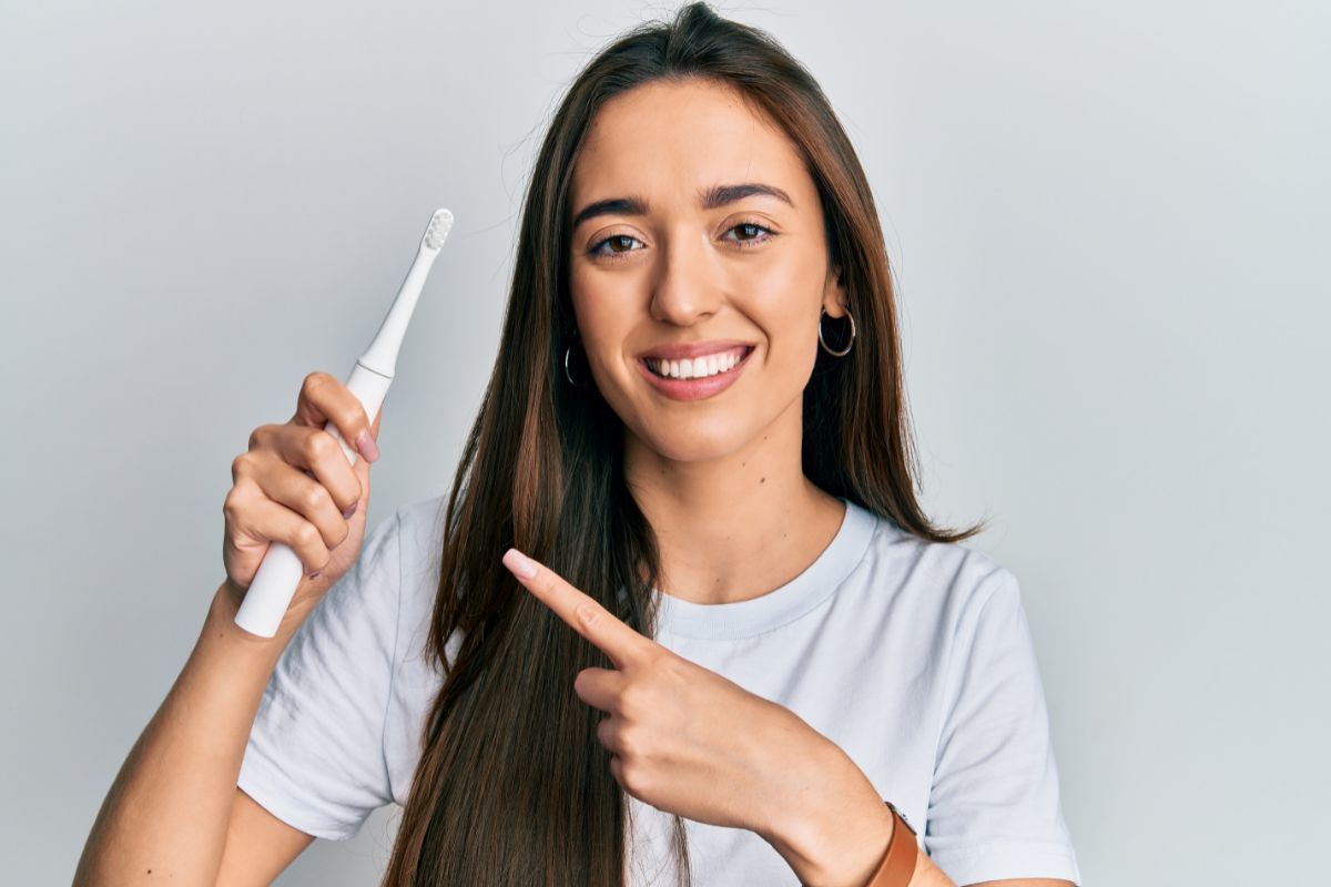Woman smiling while holding and pointing to electric toothbrush against neutral background.
