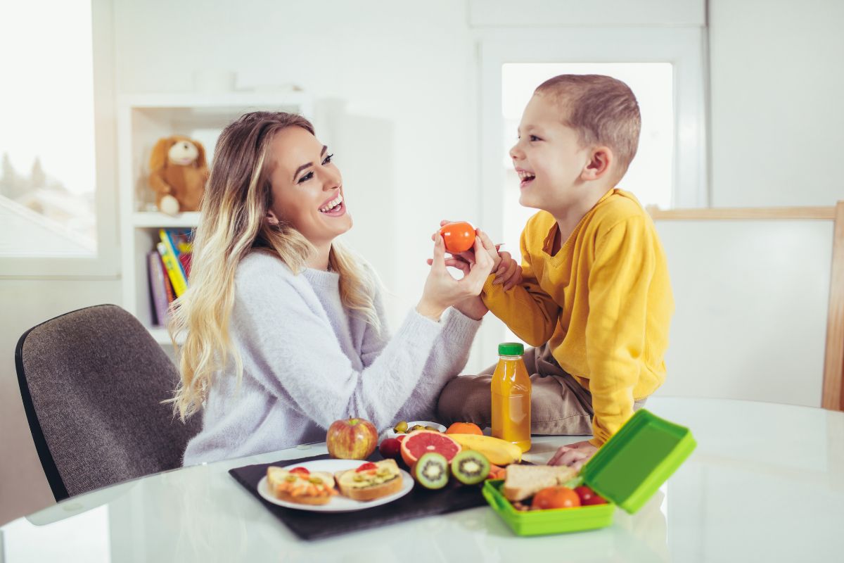 Mother and child eating healthy fruits and vegetables at kitchen table together.