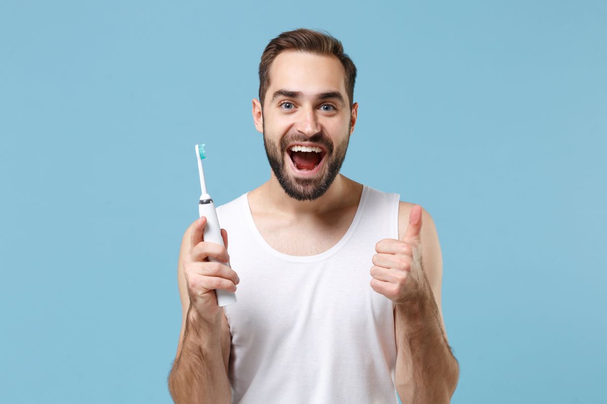 Man holding electric toothbrush giving thumbs up against blue background, promoting dental care.