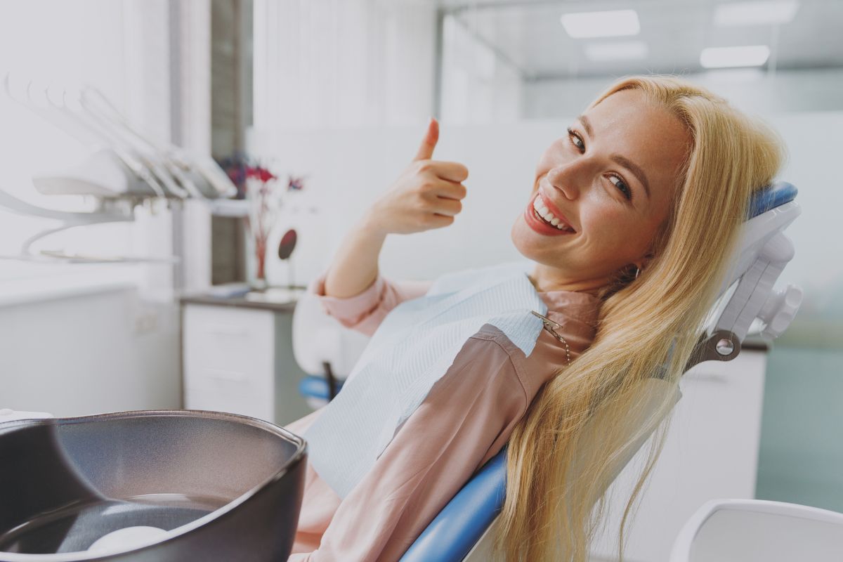 Smiling woman giving thumbs up in modern dental office chair during checkup.