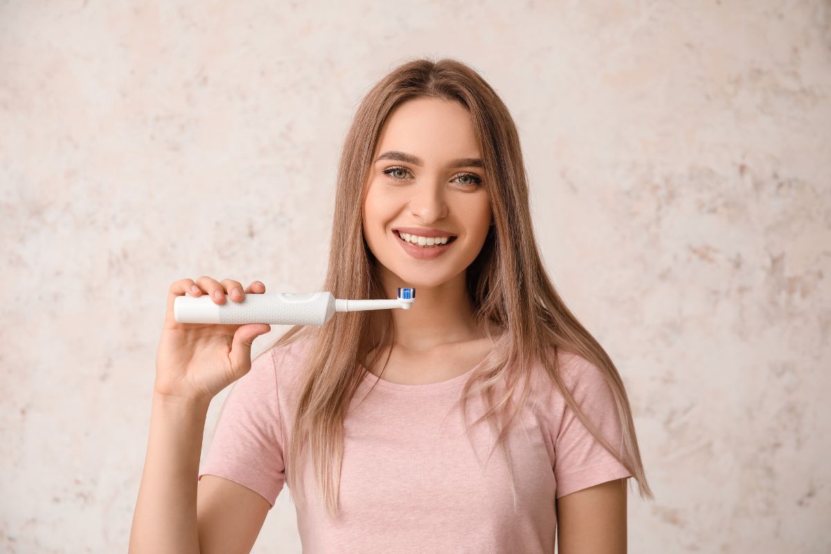 Woman in pink shirt holding toothbrush, smiling against neutral background for dental care context.