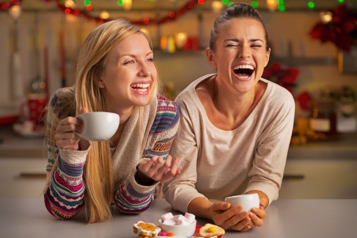 Couple laughing while holding mugs and cookies in festive holiday home setting.