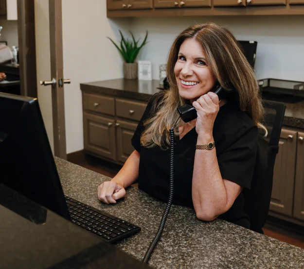 A woman seated at a desk in a dental office, smiling while speaking with a staff member